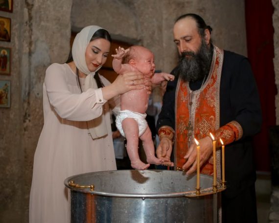 A baby being baptized by a priest and woman in a traditional church setting.